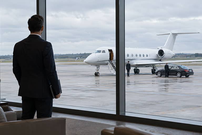 A security lead in a private terminal lounge watching a car and team prepare a private jet for departure